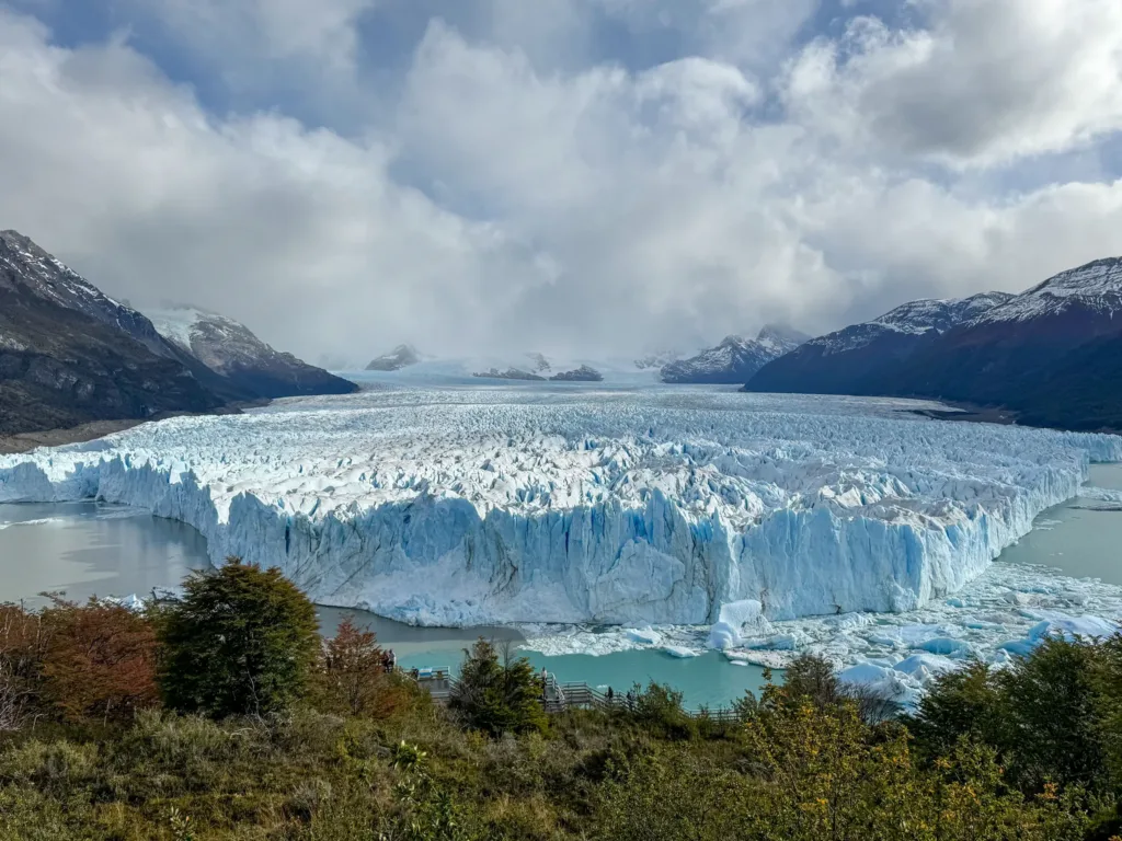 Mohutná ledová stěna ledovce Perito Moreno nad tyrkysovým jezerem, v pozadí horské vrcholy a zatažená obloha.