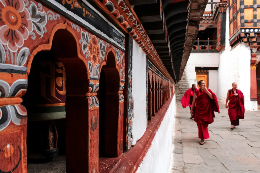 Buddhističtí mniši v červených rouchách procházejí nádvořím tradičního bhútánského kláštera s malovanými dřevěnými arkádami.
