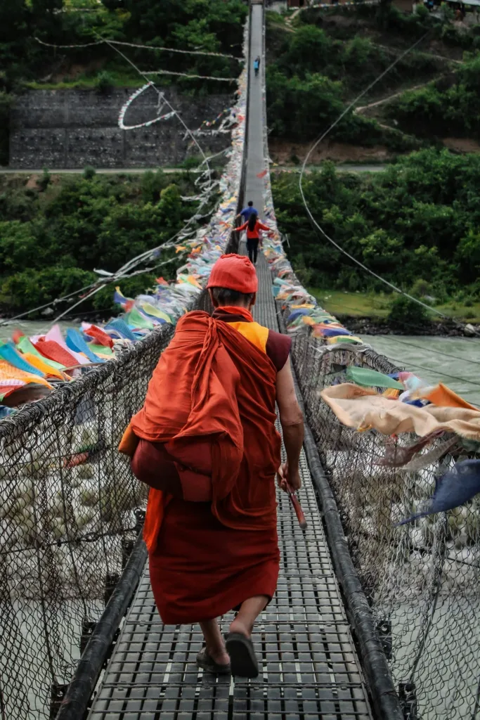 Buddhistický mnich v červeném rouchu přechází úzký visutý most ozdobený barevnými modlitebními praporky nad řekou.