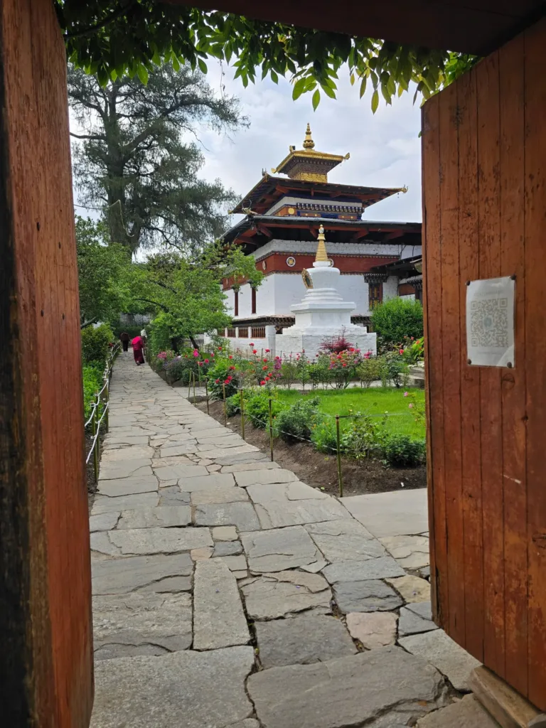 Pohled otevřenou dřevěnou branou na kamennou cestu, zahradu se stúpou a buddhistický chrám.