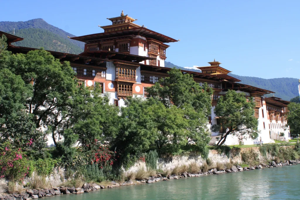 Pevnostní klášter Punakha Dzong s tradiční bhútánskou architekturou u řeky, v pozadí zalesněné hory.