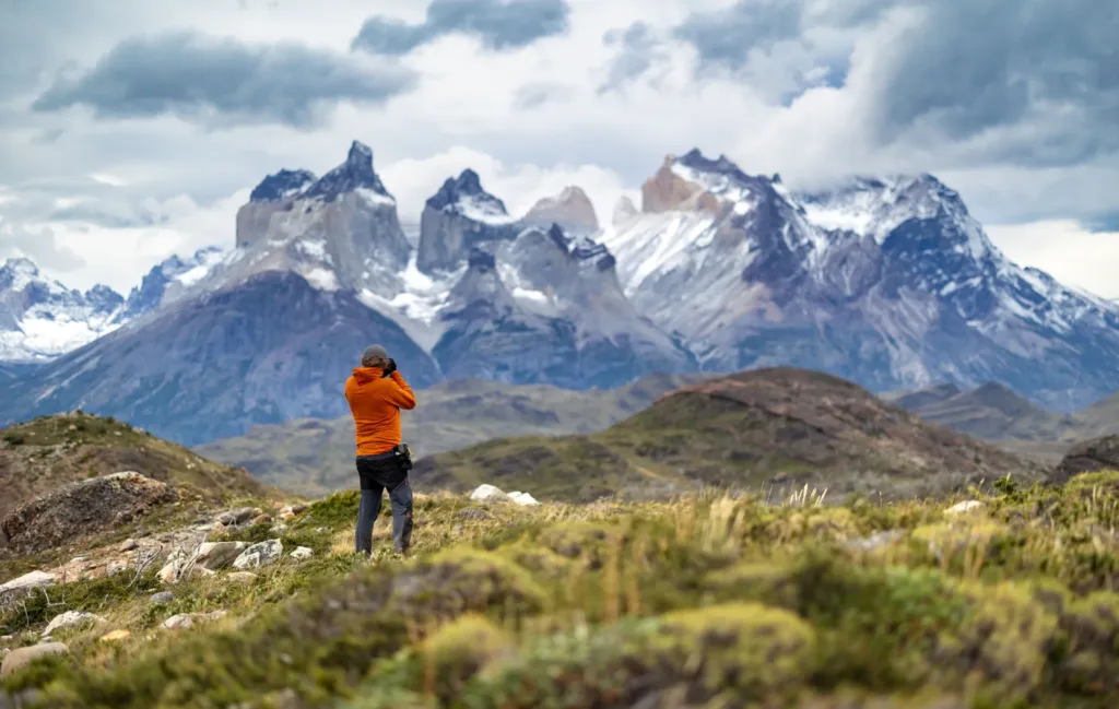 Turista v oranžové bundě fotografuje rozeklané vrcholy Cuernos del Paine v Patagonii.
