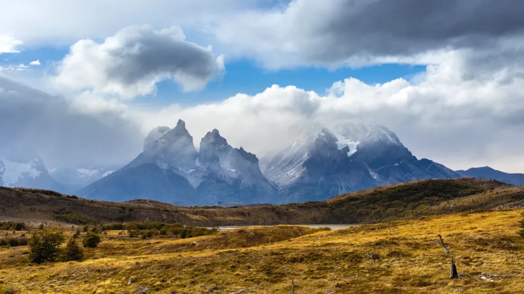 Zubaté vrcholy Cuernos del Paine vystupující nad travnatou krajinou pod dramatickými mraky.