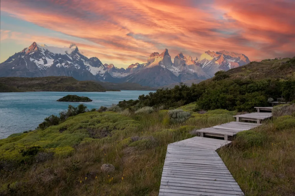 Dřevěný chodník vede zelenou krajinou podél jezera k zasněženým štítům národního parku Torres del Paine při západu slunce.