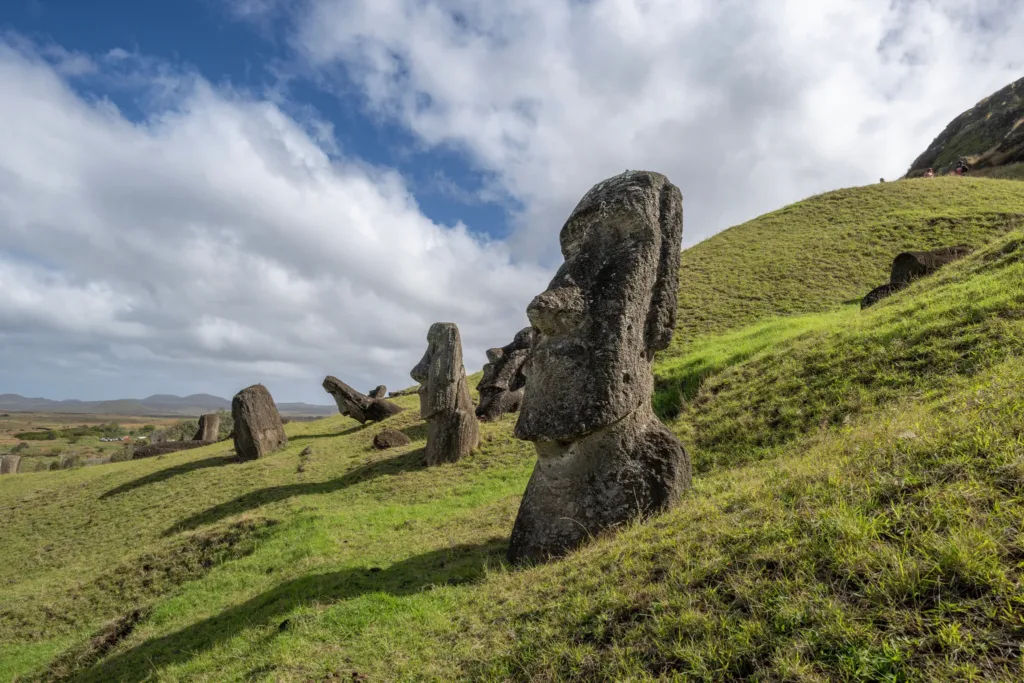 Kamenné sochy moai stojící na zeleném svahu pod zataženou oblohou.