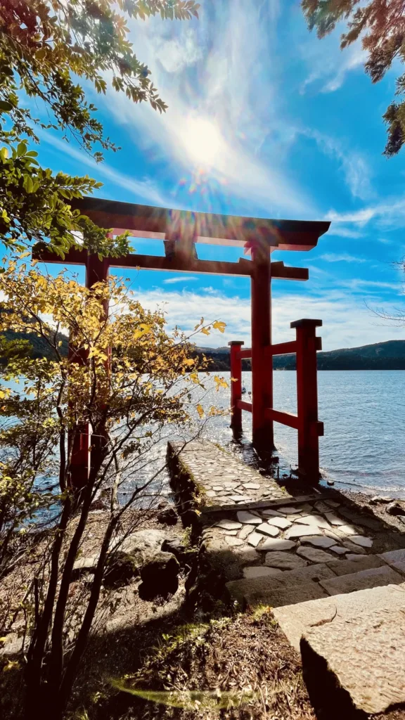 Červená brána torii stojící na břehu klidného jezera pod modrou oblohou, obklopená podzimním listím.