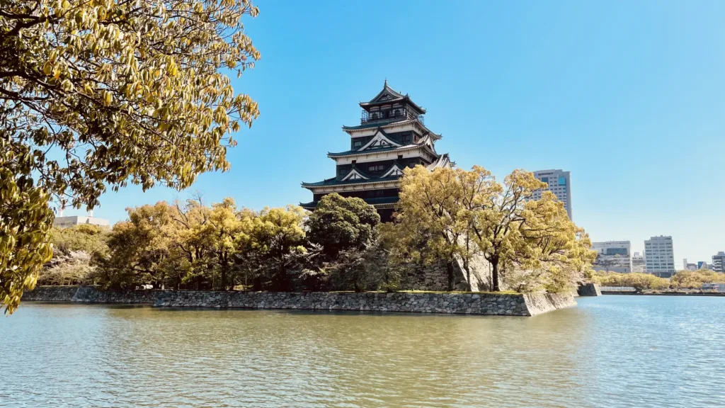 Hiroshimský Hrad obklopený stromy s podzimním zbarvením u vodního příkopu, v pozadí moderní budovy.