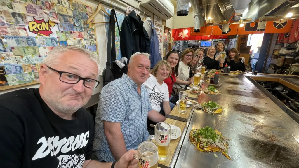 Skupina turistů sedí u dlouhého pultu v malé restauraci, před nimi jsou talíře s jídlem a sklenice piva.
