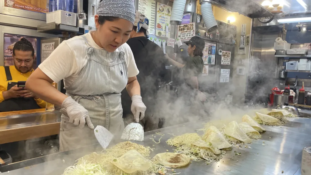 Kuchařka v otevřené restauraci připravuje na velké plotně porce japonských palačinek se zelím a nudlemi.