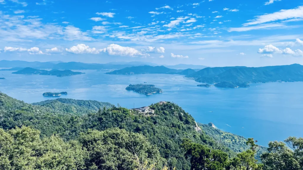 Pohled zalesněným svahem na rozsáhlé modré jezero s ostrovy a horami na obzoru.