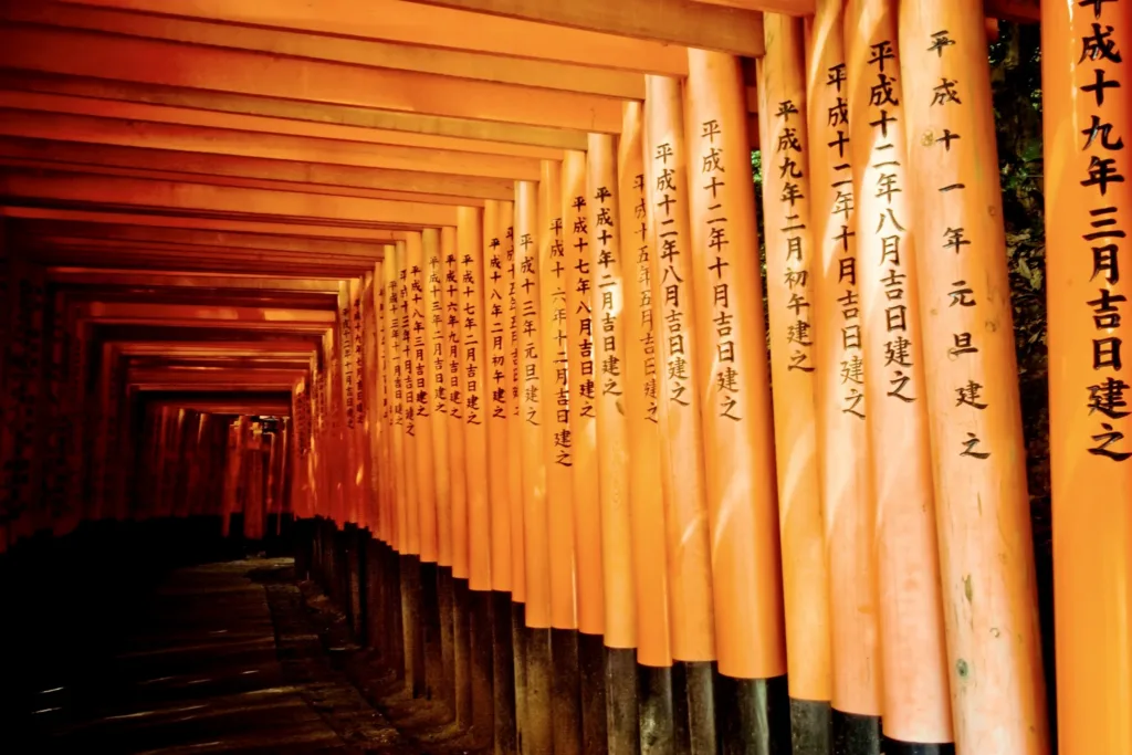 Řada oranžových bran torii s černými japonskými nápisy tvoří krytou stezku ve svatyni Fušimi Inari.