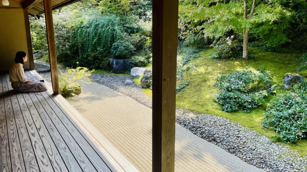 Žena sedící na dřevěné verandě s výhledem do upravené japonské zahrady s mechem, kameny a keři.