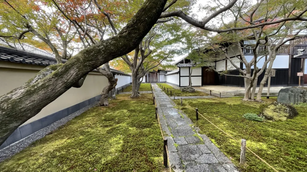Mechem porostlá zahrada s kamennou stezkou mezi tradičními japonskými budovami a podzimními stromy.