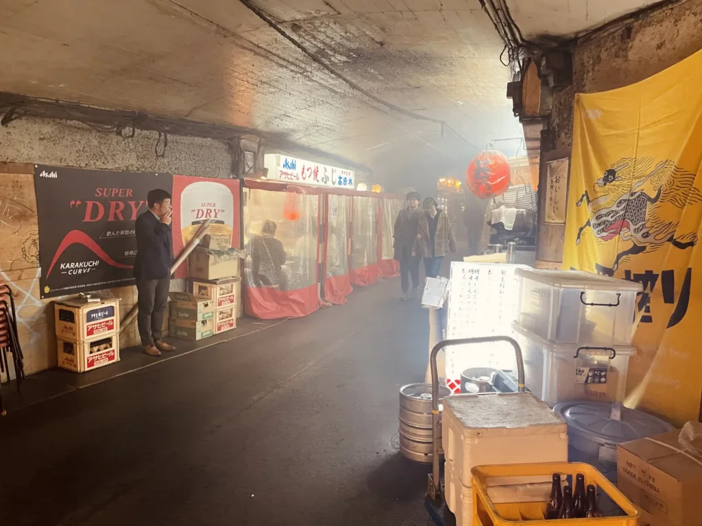 Úzká krytá ulička s malými stánky, sudy od piva a lidmi v kouři z grilování.