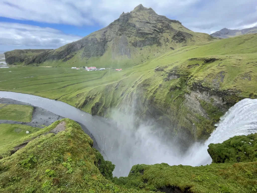 Pohled shora na vodopád Skógafoss, řeku v zeleném údolí a strmé islandské hory.