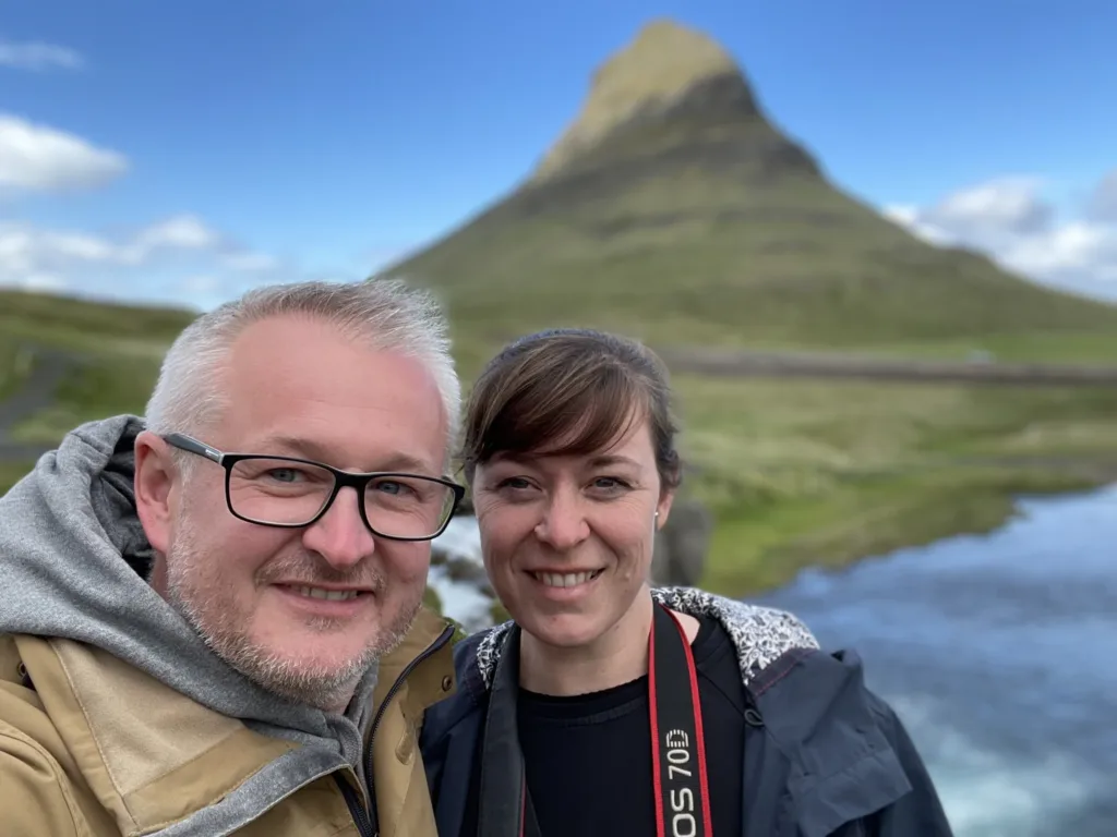 Dvojice cestovatelů pózuje u vody s horou Kirkjufell v pozadí na Islandu.