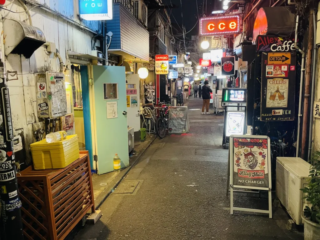 Úzká noční ulička v tokijské čtvrti Golden Gai s neonovými nápisy, malými bary a zaparkovanými koly.