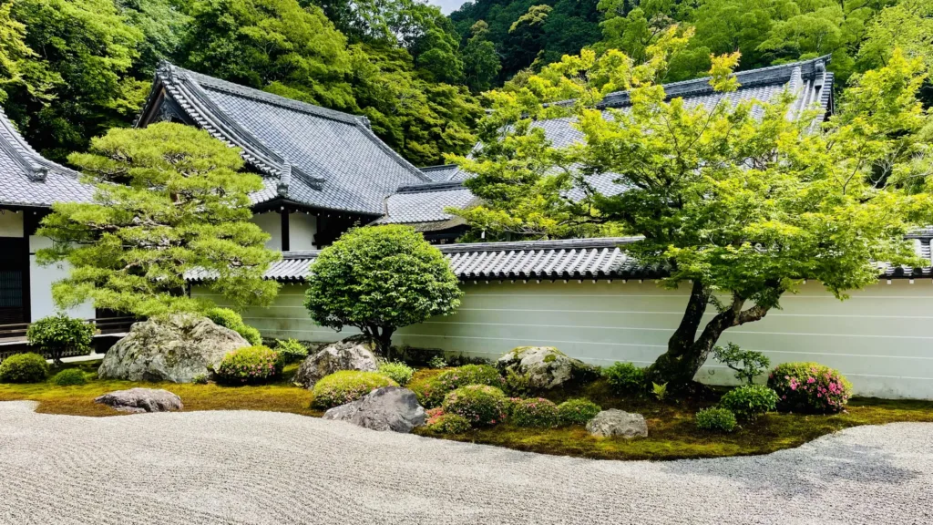 Tradiční japonská chrámová budova se šedou taškovou střechou, upravenou zahradou, kameny a štěrkovou plochou.