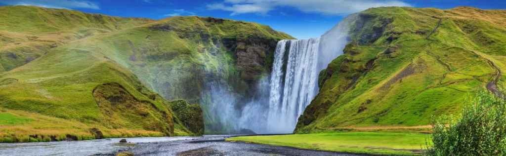 Vodopád Skógafoss padající mezi zelenými svahy na jihu Islandu, v popředí je vidět duha.