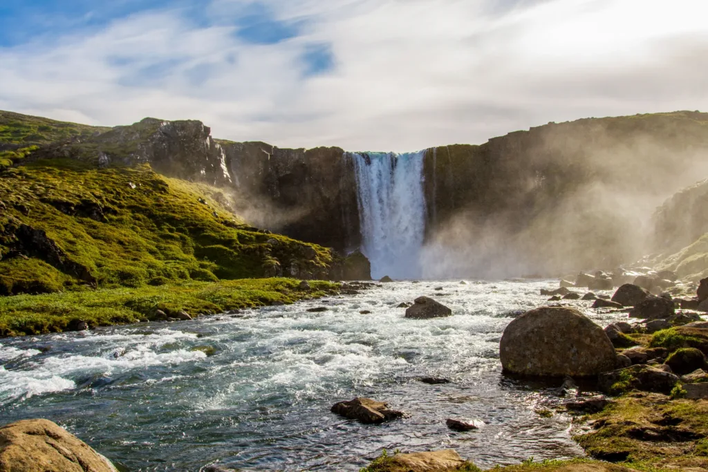 Vodopád Öxarárfoss padající z tmavých skal do kamenité řeky v zeleném islandském údolí.