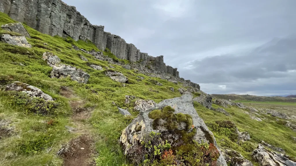 Turistická stezka vede zeleným svahem pod vysokou skalní stěnou, mezi kameny porostlými mechem.
