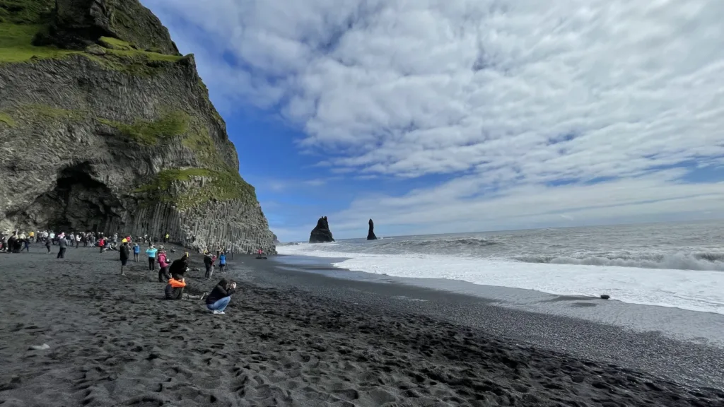 Návštěvníci na černé sopečné pláži pod strmým útesem, v moři vystupují skalní věže Reynisdrangar.