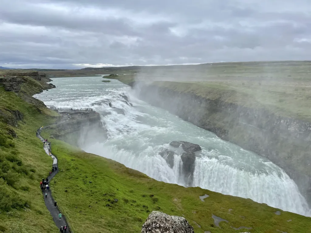 Pohled na mohutný dvoustupňový vodopád Gullfoss s tyrkysovou řekou, zelenými skalnatými břehy a pěší stezkou.