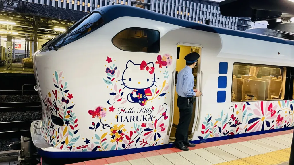 Bílý vlak Hello Kitty Haruka s květinovým motivem stojí u nástupiště, u otevřených dveří je průvodčí.