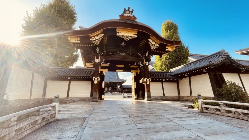 Zdobená dřevěná brána japonského chrámu mezi bílými zdmi, osvětlená nízkým sluncem.