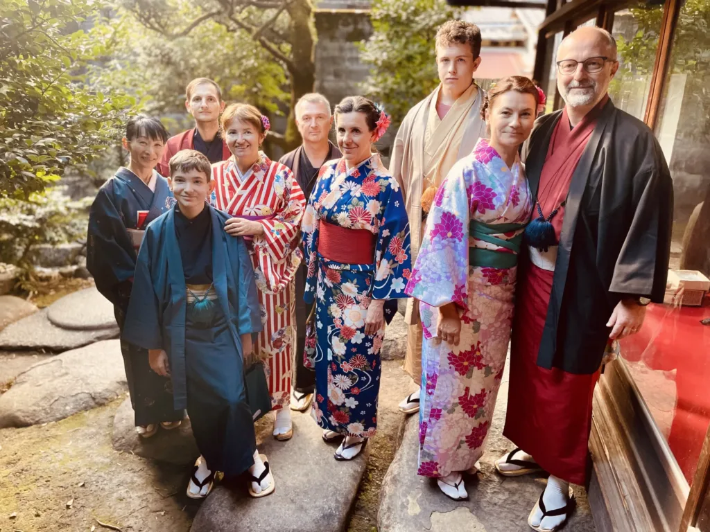 Skupina dospělých a chlapec v barevných japonských juketách stojí na kamenné cestě u dřevěné budovy.
