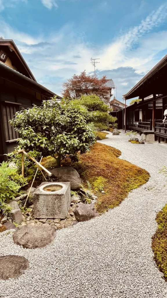 Štěrková zahrada s nízkými keři a kamennou nádobou mezi dřevěnými domy v tradičním japonském stylu.