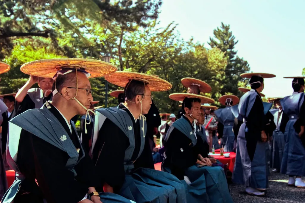 Účastníci tradiční japonské slavnosti v tmavém oděvu a širokých slaměných kloboucích sedí a stojí pod stromy.