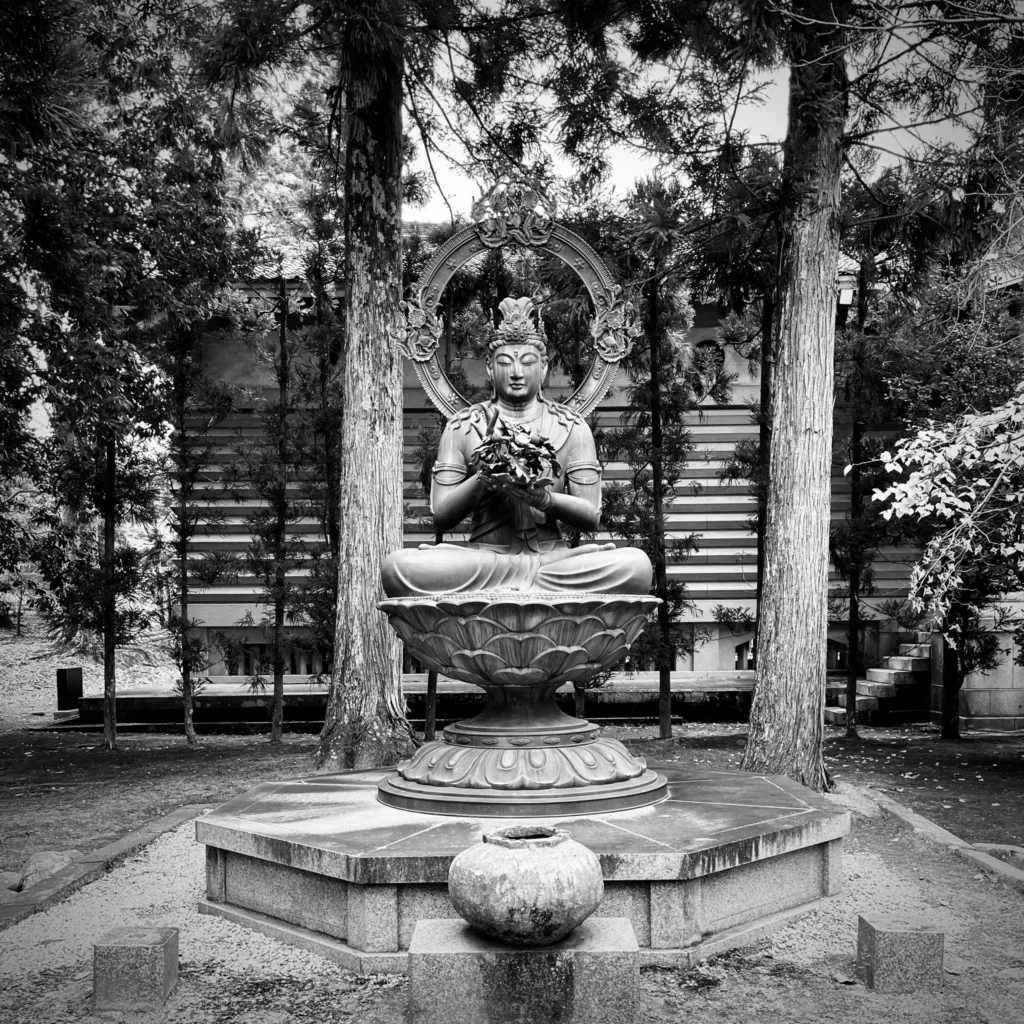 Sedící buddhistická socha na zdobeném podstavci mezi stromy v zahradě.