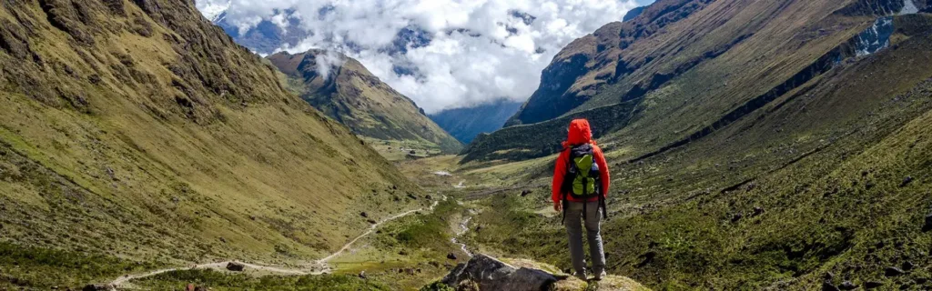 Turista s červeným batohem kráčí po stezce v zeleném horském údolí pod zataženou oblohou.