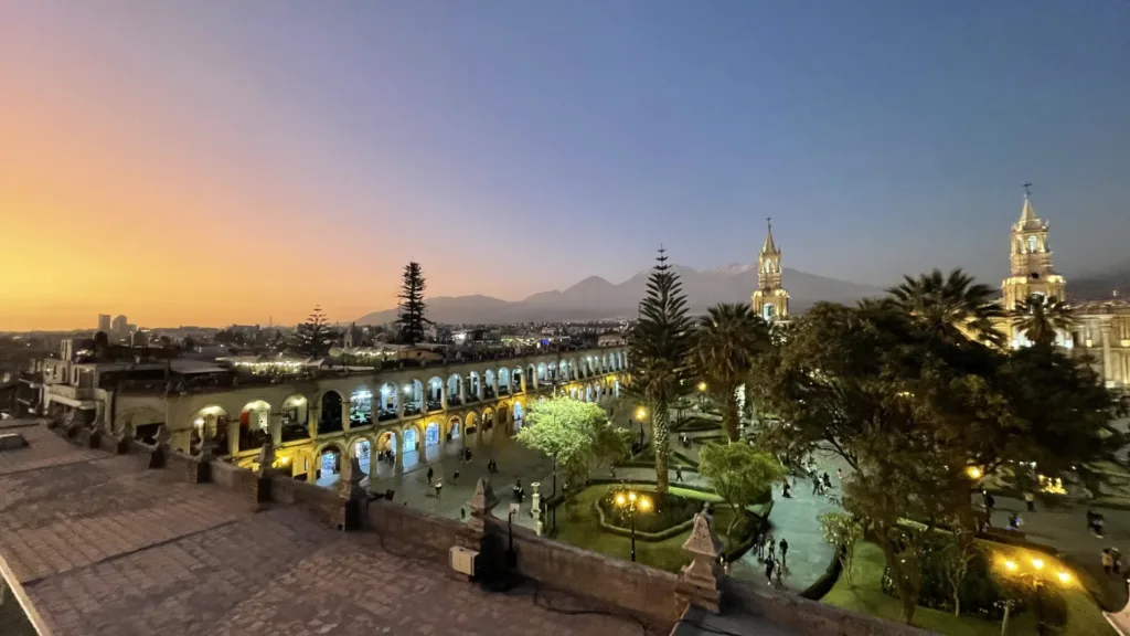 Osvětlené náměstí Plaza de Armas v Arequipě s arkádami, palmami a věžemi katedrály za soumraku.