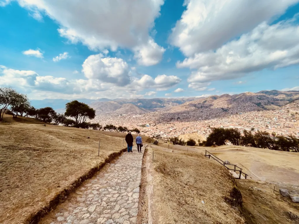 Dva lidé kráčejí po kamenné stezce nad městem Cusco, obklopeným andskými horami.