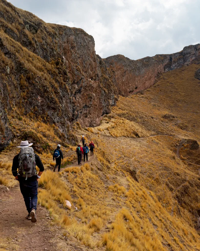 Skupina turistů stoupá po stezce mezi skalnatými svahy a vysokohorskou travinou.