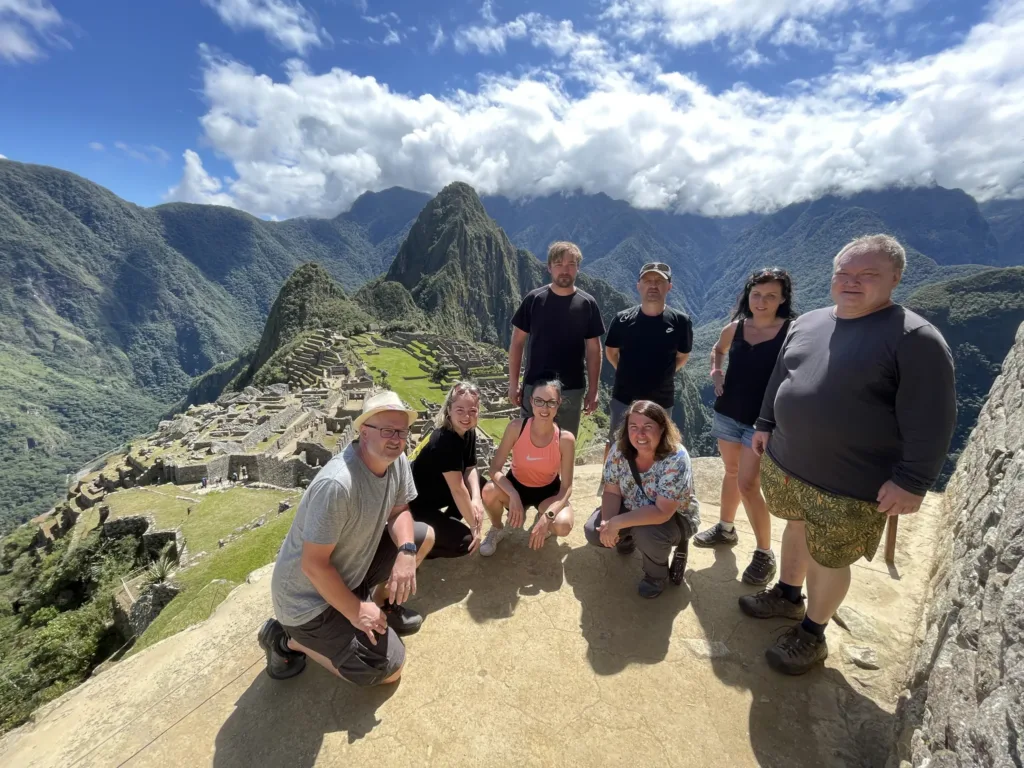 Skupina turistů pózuje na vyhlídce nad ruinami Machu Picchu a zelenými andskými horami v Peru.