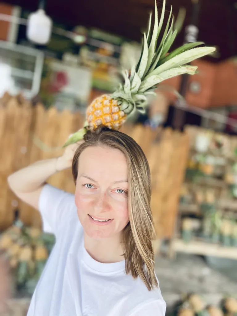 Usměvavá žena v bílém tričku drží na hlavě malý ananas před obchodem.