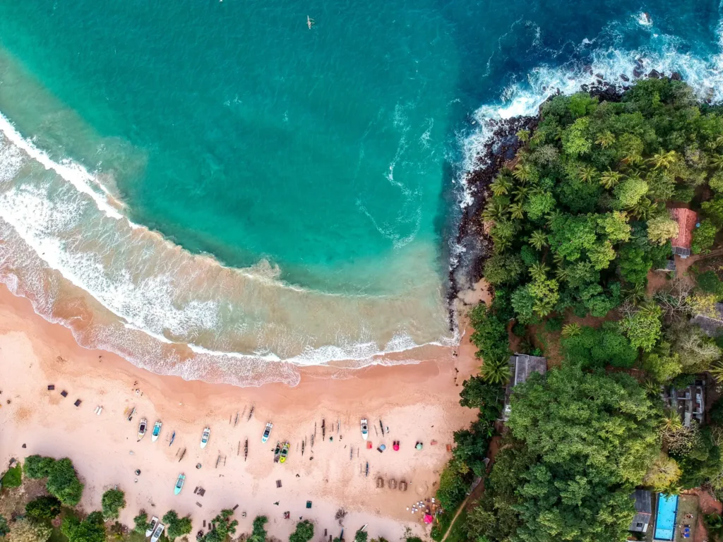 Letecký pohled na písečnou tropickou pláž s tyrkysovým mořem, malými čluny a hustou zelení u skalnatého výběžku.