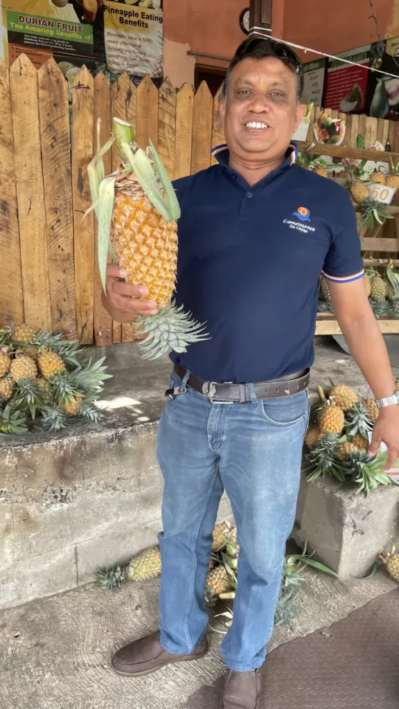 Usměvavý muž drží velký ananas před stánkem s tropickým ovocem.