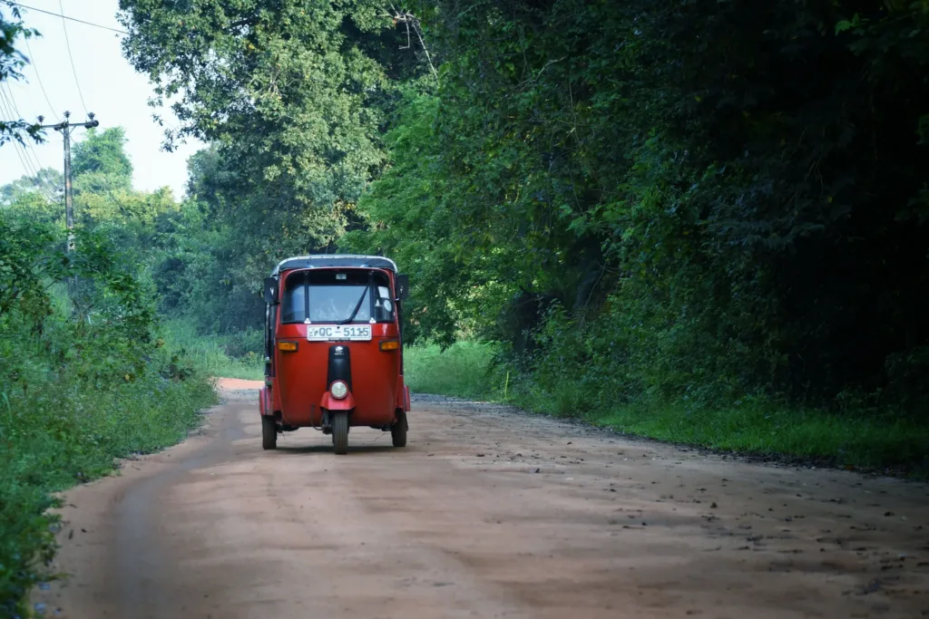 Červený tuk-tuk jede po prašné cestě lemované hustou tropickou vegetací.