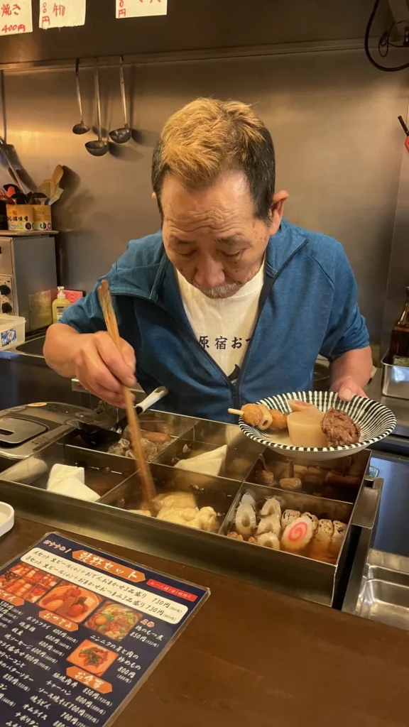 Muž v modré mikině nabírá hůlkami ingredience z hrnce s japonským pokrmem oden.