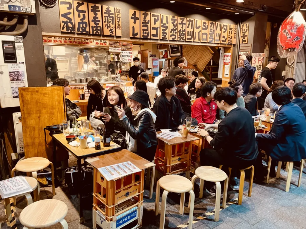 Hosté sedí u malých stolů v rušné japonské restauraci s ručně psanými jídelními lístky nad pultem.