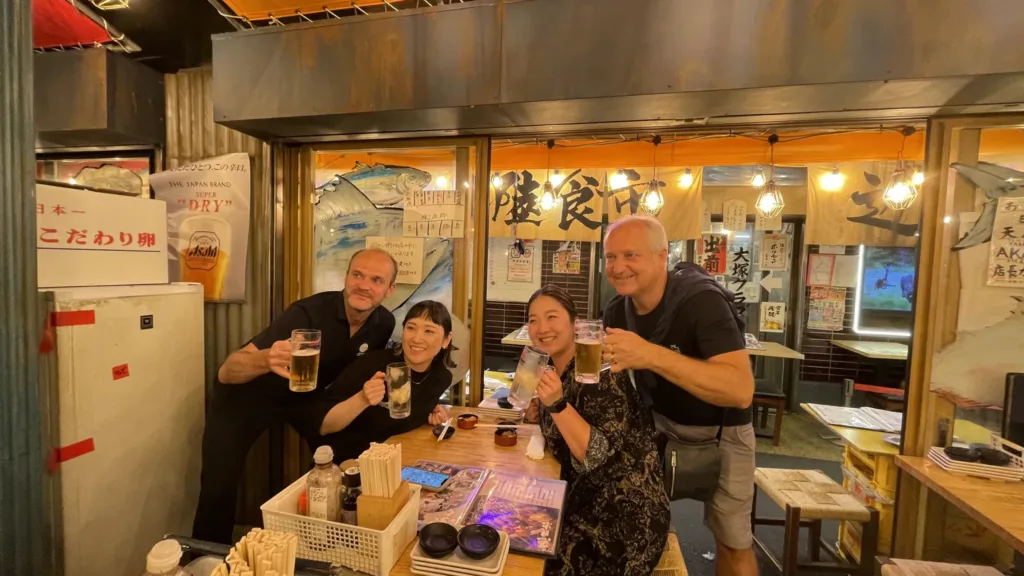 Čtyři lidé připíjejí pivem u stolu v malé japonské restauraci s lampiony a japonskými nápisy.