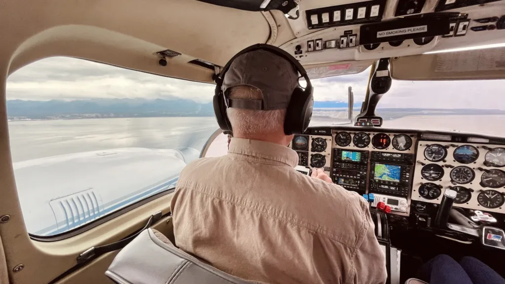 Pilot v kokpitu malého letadla při letu nad klidnou vodní hladinou, v dálce jsou vidět hory.