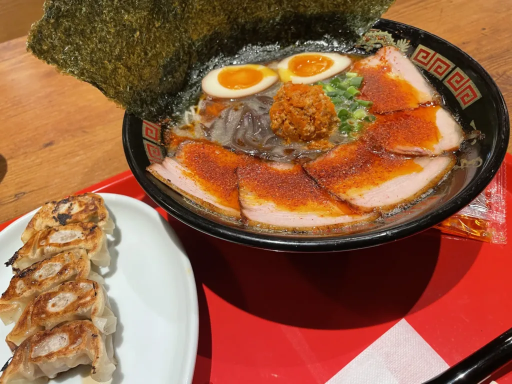 Miska ramen s plátky masa, vejcem, mořskou řasou a pálivým kořením, vedle talířek grilovaných taštiček gyoza.