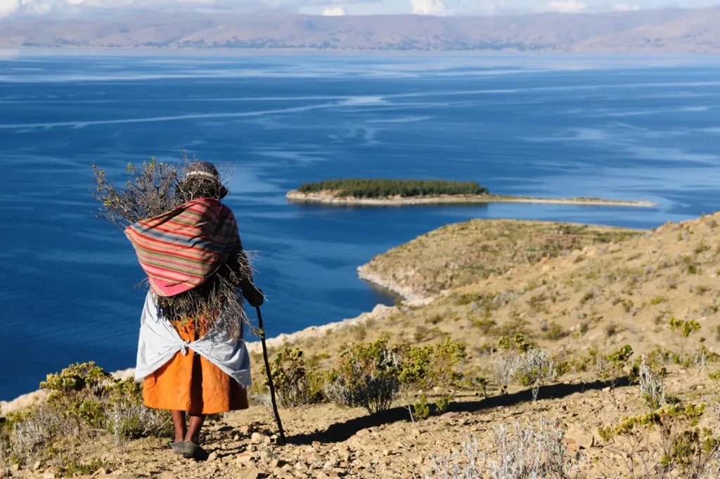 Žena v tradičním barevném oděvu stojí na suchém svahu nad modrou hladinou jezera Titicaca a hledí na malý ostrov.