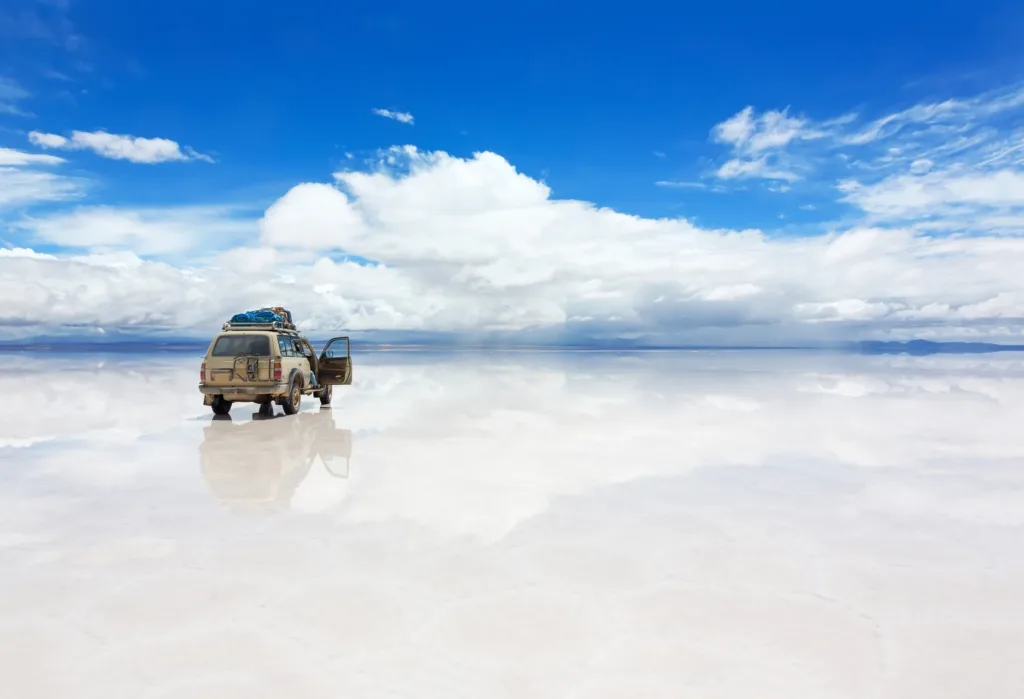 Terénní vůz stojící na lesklé solné pláni Salar de Uyuni v Bolívii pod modrou oblohou s mraky.