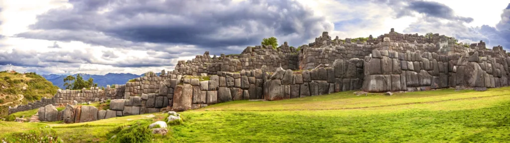 Mohutné kamenné zdi pevnosti Sacsayhuamán nad zeleným trávníkem pod zataženou oblohou